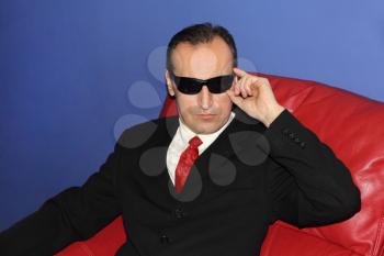 Man in black glasses sits on red sofa and looks at camera.