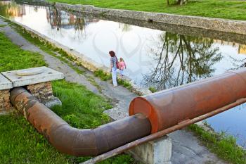 Woman and little girl going by waters bank. Large rusty metal pipe over the reservoir. View from the top