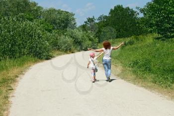 Mother and daughter walking by rural road among trees in lovely summer day