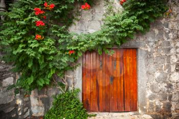 Red wooden gate in old stone wall with decorative flowers. Perast town, Montenegro