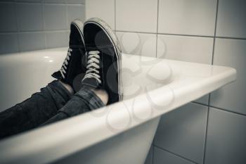 Teenager feet in black sporty sneakers lay in white bath, dark vintage tonal correction photo filter effect 