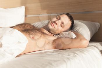 Young European man sleeping in wide bed under white blanket in morning sunlight