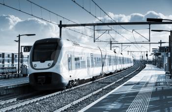 Electric passenger train goes near railway platform in Amsterdam, Netherlands. Blue toned monochrome photo