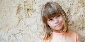 Slightly smiling Caucasian little girl, close-up outdoor portrait over stone wall