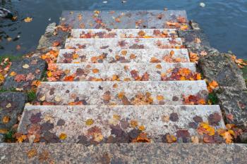 Stone stairway with red fallen leaves goes down to blue pond water in autumn park