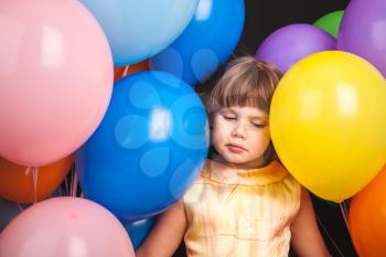 Portrait of sad Caucasian blond little girl with colorful balloons over black background