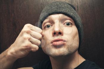 Young aggressive Caucasian man in gray hat shows fist. Closeup studio face portrait, selective focus
