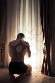 Young adult praying man sits on the knees near glowing window in dark room at night, rear view