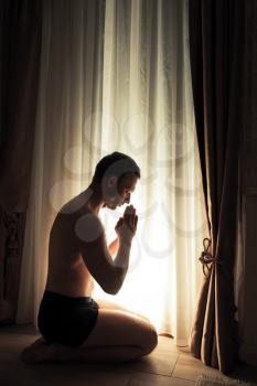 Young adult praying man sits on the knees near glowing window in dark room at night