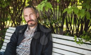 Young bearded Asian man on white bench in summer park, outdoor portrait