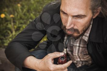 Young serious bearded man smoking pipe, close up outdoor portrait