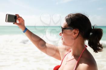 Smiling young adult Caucasian woman in bright swimsuit and sunglasses takes selfie photo on the beach of Saona island, Dominican Republic. Outdoor profile portrait