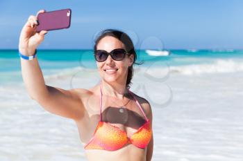 Smiling young adult Caucasian woman in bright swimsuit and sunglasses takes selfie photo on the beach of Saona island, Dominican Republic