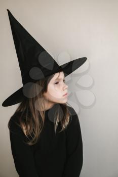Little blond girl in black witch costume stands near white wall, close-up portrait
