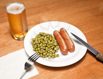Daily food theme. Sausages with mustard, green peas and glass of beer on wooden table