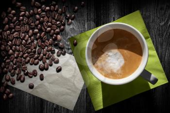 Cup of cappuccino coffee, old paper sheet, green napkin and beans on black wooden table, top view