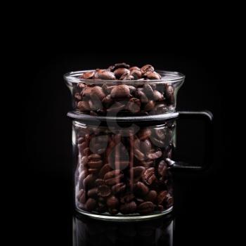 Mug made of glass is full of coffee beans on black background