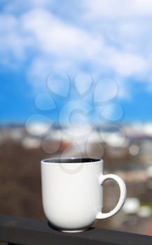 White cup stands on balcony railing above blurred cityscape with steam looks like clouds on blue sky