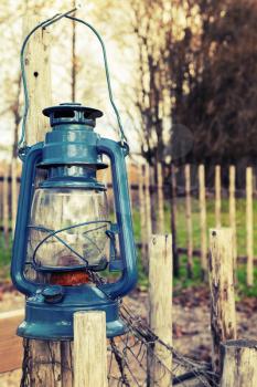 Old blue kerosene lamp hangs on wooden outdoor fence, vintage toned photo with filter effect