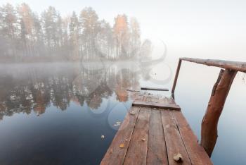 Small wooden pier on still lake in cold foggy morning