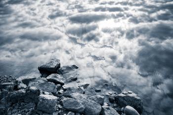 Still lake coast, coastal stones and cloudy sky reflection in water, monochrome photo