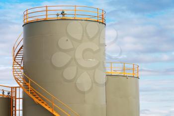 Gray cylindrical fuel tanks with yellow railings above blue cloudy sky