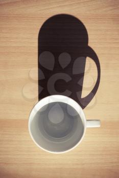 Empty white ceramic coffee cup on wooden table background with strong dark shadow from the sunlight. Photo filter effect