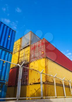 Colorful Industrial cargo containers are stacked under blue cloudy sky behind metal fence with barbed wire. Vertical photo