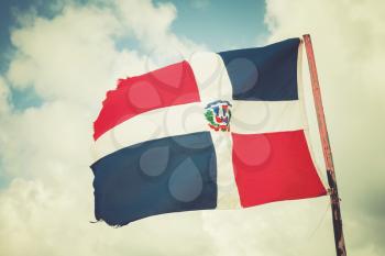 Flag of Dominican republic is waving on wind over blue cloudy sky background, old style photo filter effect with warm tonal correction