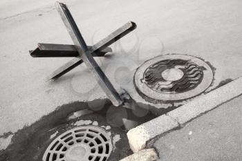 Black steel street barrier in shape of anti-tank Czech hedgehog obstacle defense stands on asphalt urban road near sewer manholes
