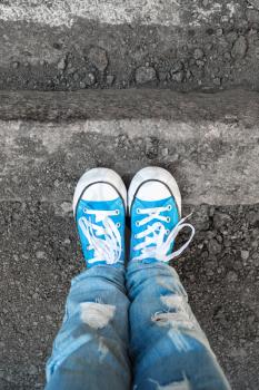 Teenager feet in jeans and blue shoes stand on the street edge. Close-up photo with selective focus and shallow DOF