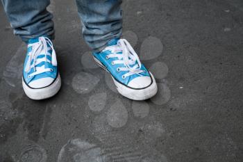 Blue sneakers, teenager feet in gumshoes stand on dark gray asphalt. Closeup photo with selective focus and shallow DOF
