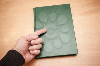 Male hand with pointing finger shows on book with empty dark green leather cover laying on wooden desk
