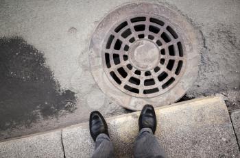Urbanite man in black new shining leather shoes standing on the roadside near rusty sewer manhole