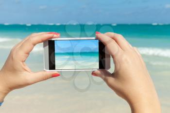 Woman holds smart phone in hands for taking photo on a summer beach in Dominican republic