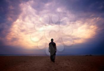 Man goes to the sea on dark sand beach under beautiful colorful cloudy sky