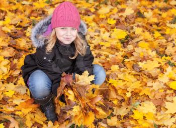 Little blond girl seat on the park ground with yellow autumn leaves. Outdoor smiling portrait