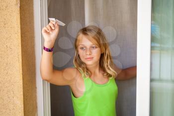 Little blond Caucasian girl with small paper plane in window, outdoor close-up portrait