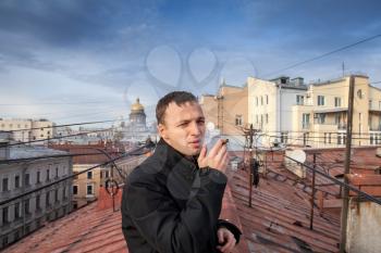 Young Caucasian man smokes cigar on the roof in central part of Saint-Petersburg, Russia