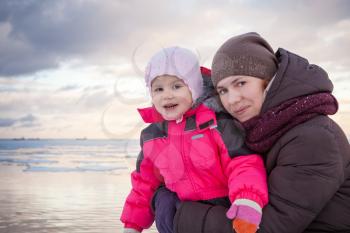 Caucasian family outdoor portrait on winter sea coast, young mother hugs her baby girl in pink jacket
