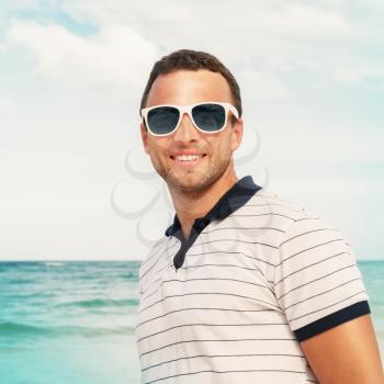 Outdoor portrait of young smiling Caucasian man in white sunglasses on summer sea coast. Vintage toned photo with Instagram style photo filter effect