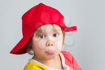 Studio portrait of funny confused baby girl in red baseball cap over gray wall background