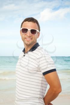 Outdoor portrait of young smiling Caucasian man standing with white sunglasses on summer sea coast. Vintage toned photo with style filter effect