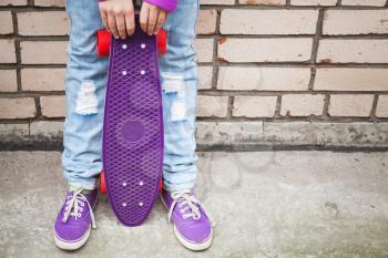 Teenage girl in jeans and gumshoes holds skateboard near by gray urban brick wall