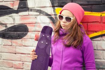 Blond beautiful teenage girl holds skateboard near by urban wall with colorful graffiti