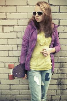 Blond teenage girl in jeans and sunglasses holds skateboard near gray urban brick wall, vintage cold green tonal correction, old style photo filter effect