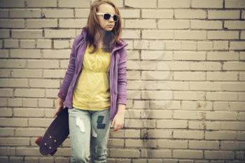 Blond teenage girl in jeans and sunglasses holds skateboard near gray urban brick wall, vintage tonal correction, old style photo filter effect