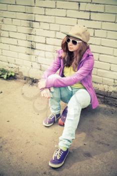 Beautiful teenage girl in cap and sunglasses sits on a skateboard near brick wall, vertical photo with warm retro tonal correction effect, instagram old style filter