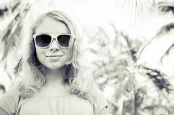 Blond smiling girl teenager in sunglasses, closeup outdoor summer portrait with palms on a background, monochrome retro style photo filter effect