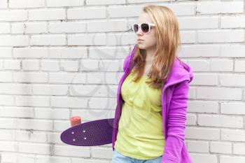 Blond teenage girl in sunglasses holds skateboard near white urban brick wall